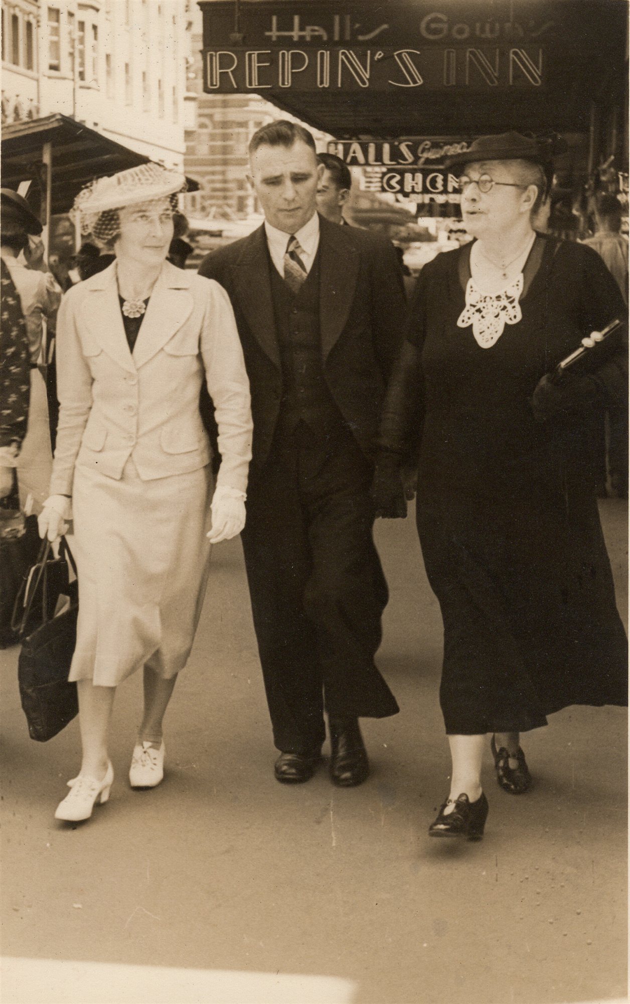 Two women and a man walk together on a city footpath past a shopfront lit with neon signs reading "Hall’s Gowns" and "Repin’s Inn".