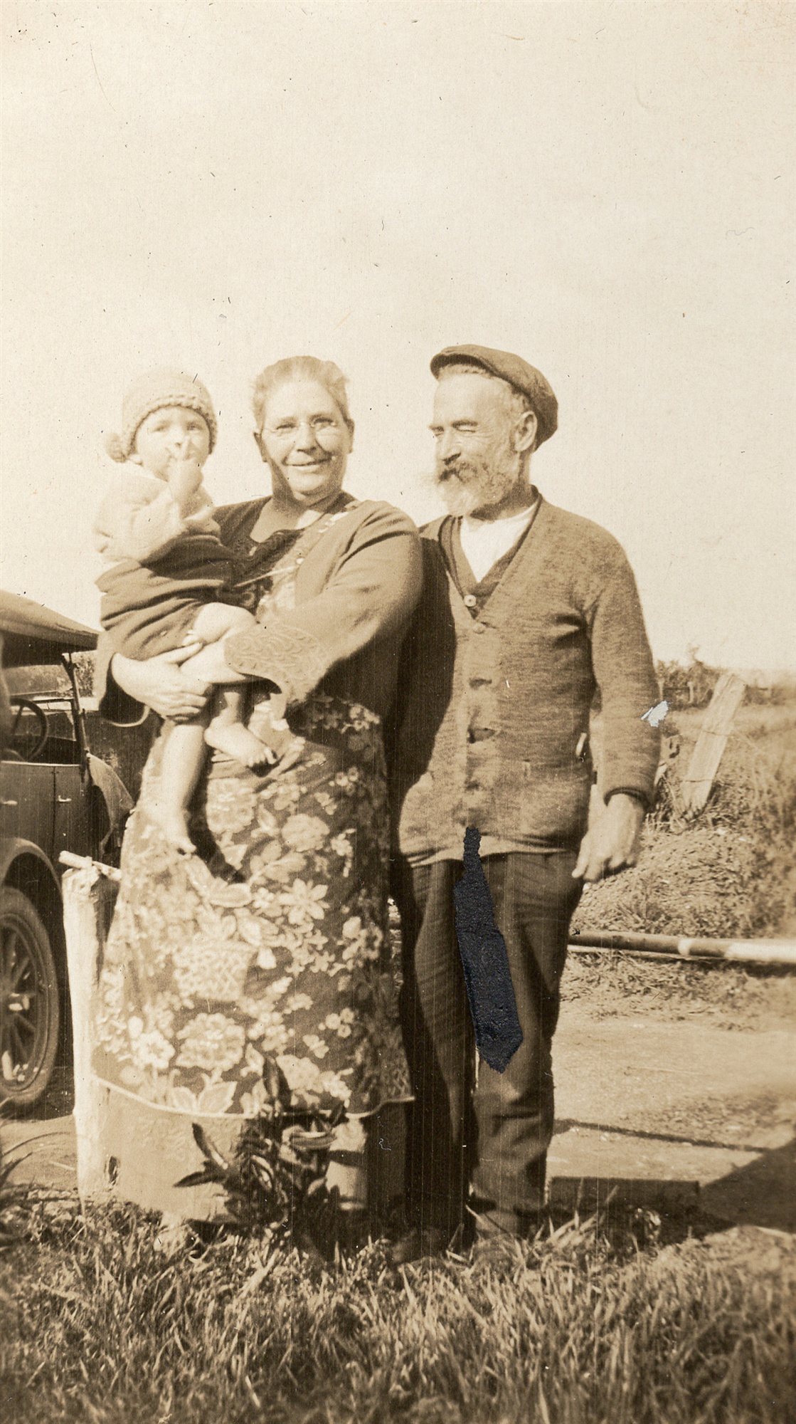 An elderly woman holds a small child in a knitted bonnet, standing beside an elderly man in a flat cap and cardigan, a motor car parked behind them.
