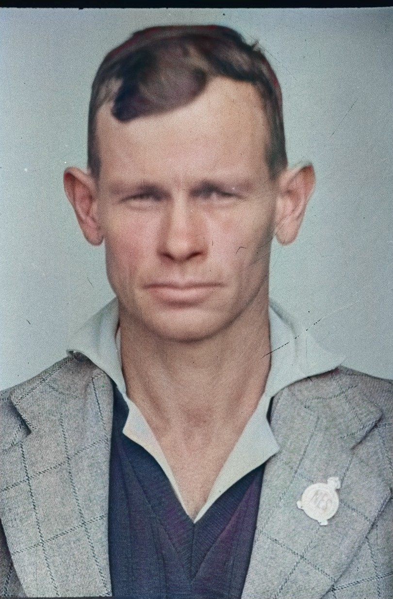 Head-and-shoulders portrait of a lean man with close-cropped hair, in a checked jacket over an open-necked shirt, a small round badge on the lapel — hand-coloured.