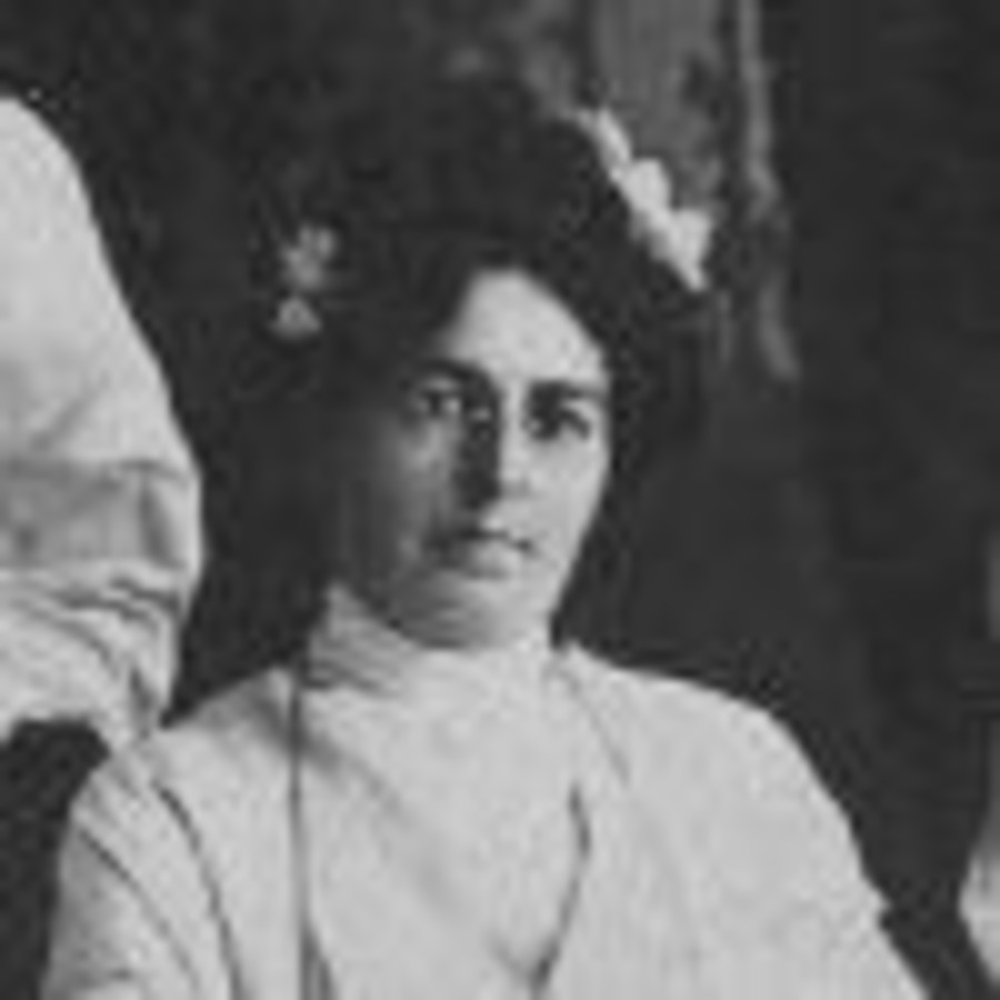 Head-and-shoulders detail of a dark-haired woman in a high-necked white blouse, a flower pinned in her hair.