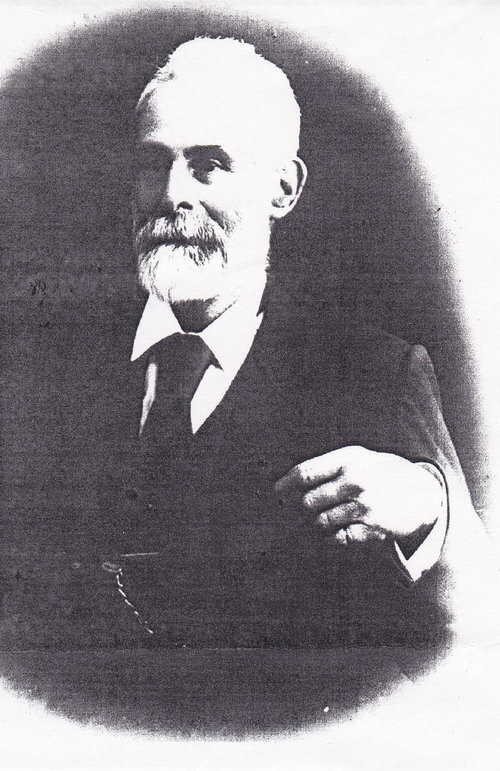 An elderly white-bearded man in a dark three-piece suit and watch chain, photographed side-on, one hand raised at his chest.