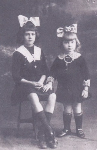 A young girl seated on a stool beside her small sister standing, both in dark dresses with large white bows in their hair, studio portrait.