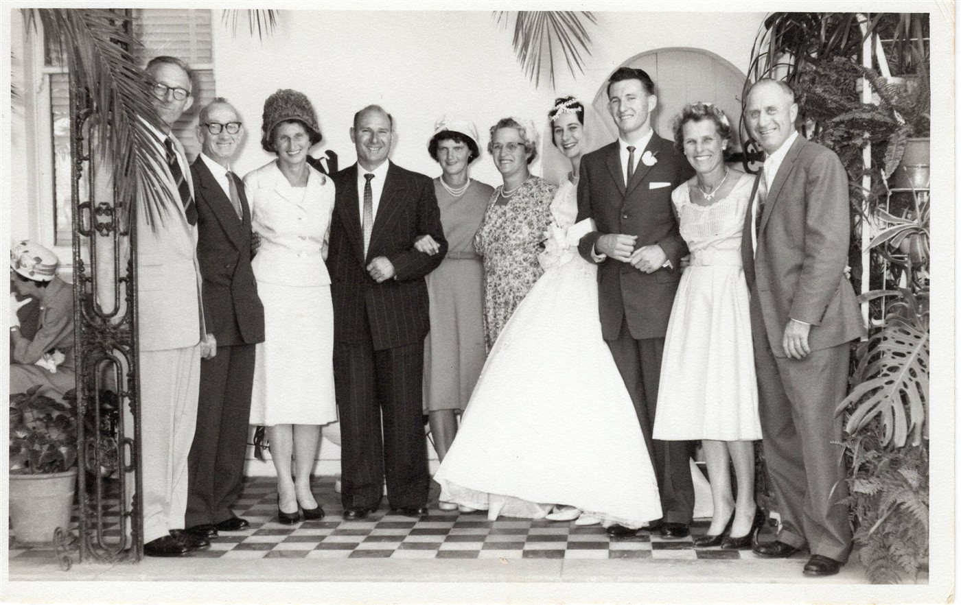 A black-and-white wedding photograph of ten people standing together outside a church, the bride in a full white gown at centre.