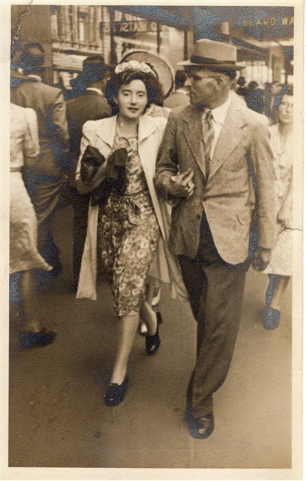 A man and woman in 1940s dress walking arm in arm along a busy city street.