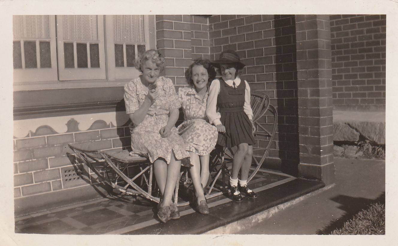 Three women and a girl seated and standing on a verandah bench: an older woman, a younger woman beside her, and a girl in school uniform standing at the end.