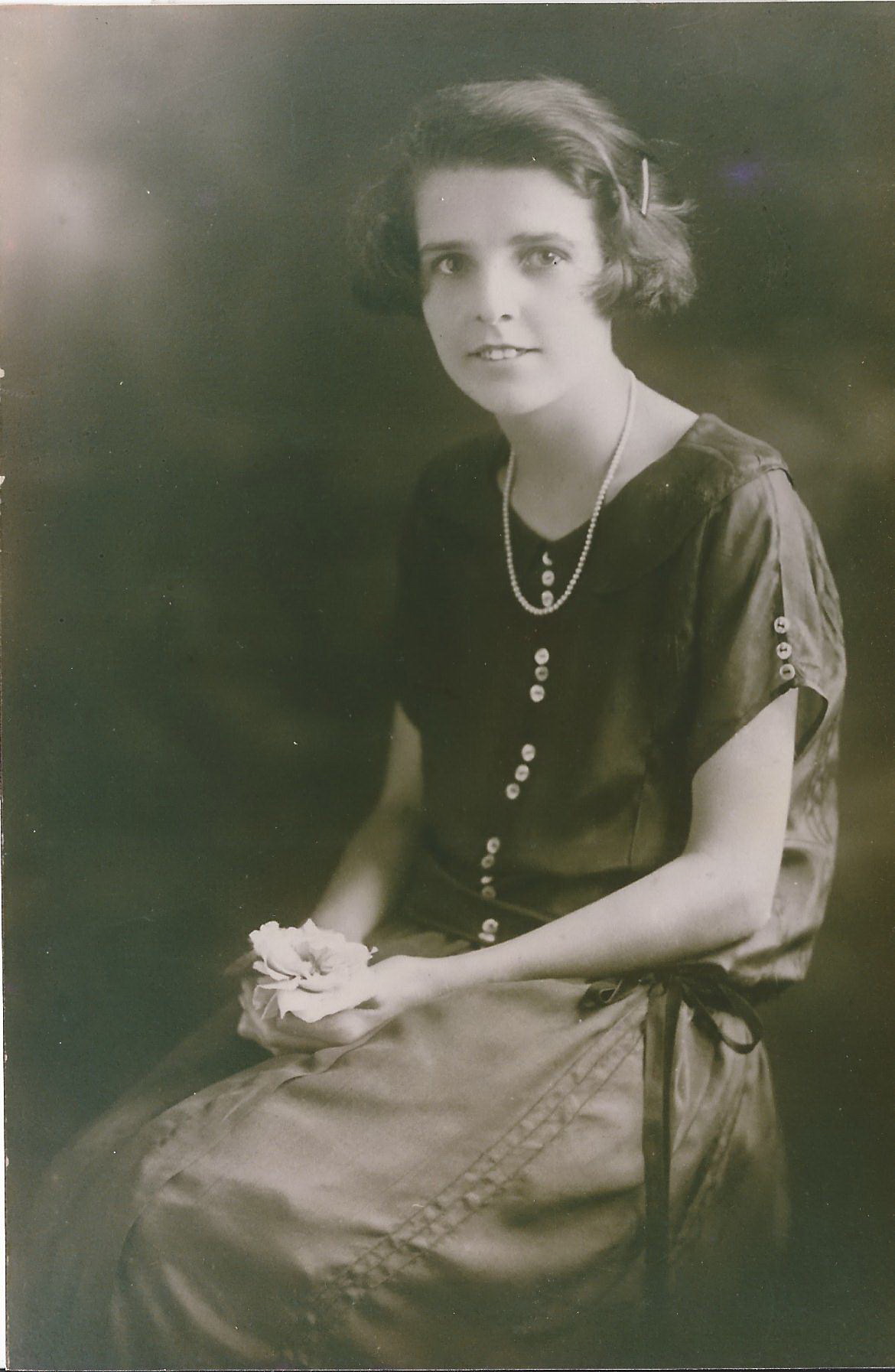 A young woman with short waved hair, seated in a studio portrait, wearing a dark dress and a long pearl necklace, holding a white rose.