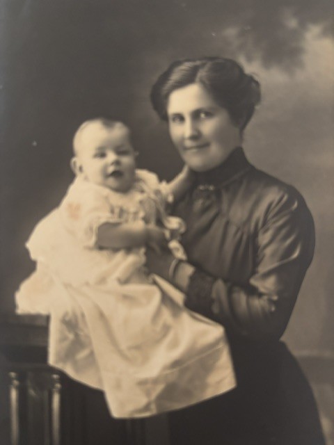 A sepia studio portrait of a young woman in a dark high-necked dress holding an infant in a long white christening gown.