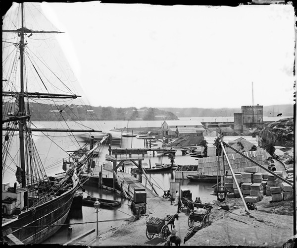 Black-and-white photograph looking across Circular Quay from Dawes Battery toward Campbell’s Wharf, Sydney, September 1873: a tall-masted sailing ship in the foreground, small boats and lighters moored along the quay, laden carts on the wharf road, and the castellated tower of Fort Macquarie in the distance.