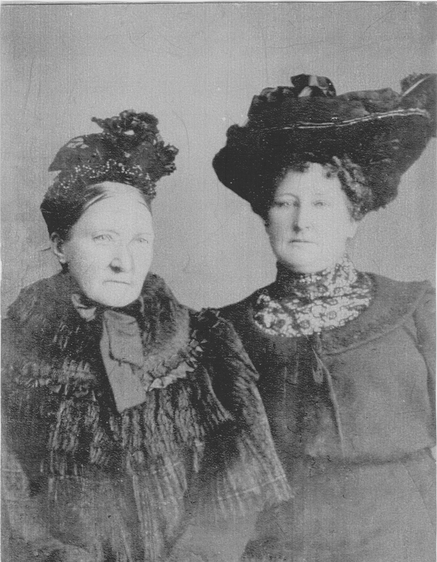 A formal studio double portrait of two women in dark, fur-trimmed coats and elaborate wide-brimmed feathered hats, in the style of the early 1900s.
