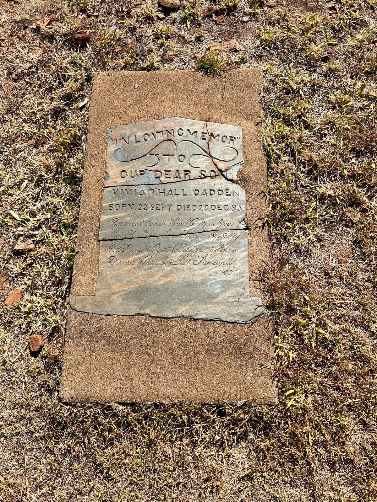 A broken slate headstone set flat in a concrete surround on dry grassy ground at the Old Halls Creek Cemetery.