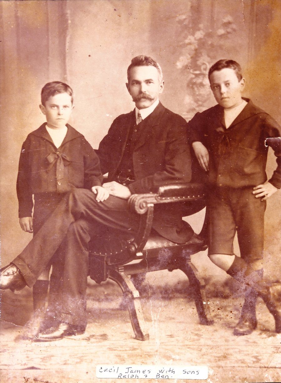 Sepia studio portrait of a moustached man seated in a carved chair with a boy standing at either side, both in sailor collars; a handwritten caption is pasted below.