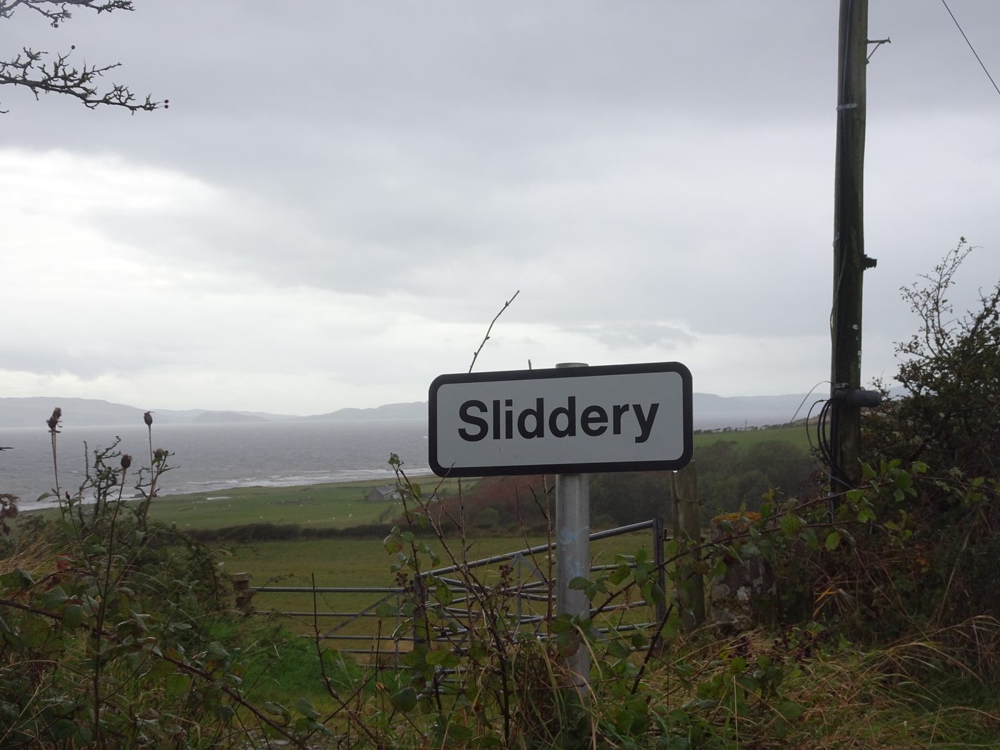 A road sign reading Sliddery beside a gate, with green fields running down to the sea under a grey sky.