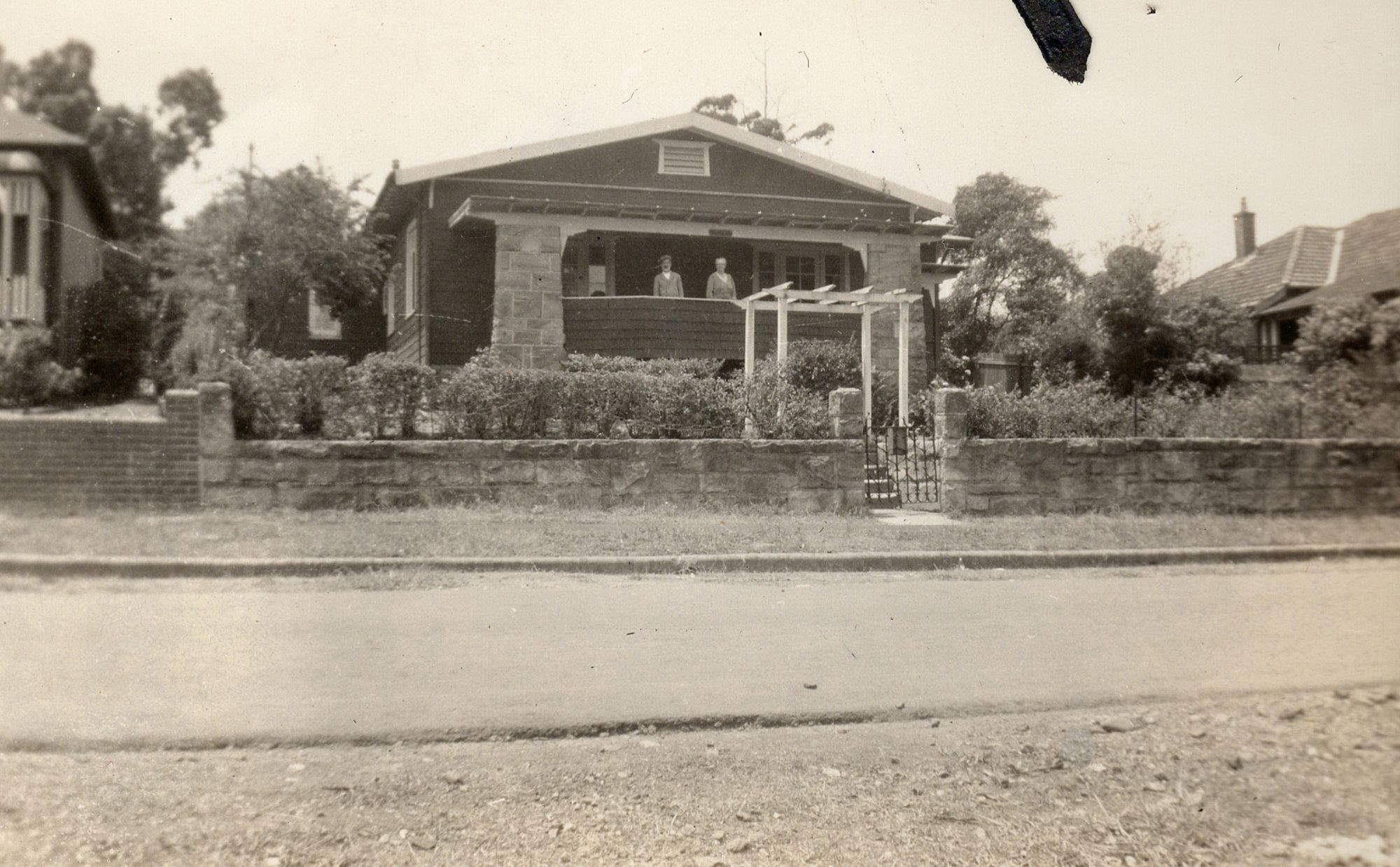 Photograph of a brick California bungalow behind a low sandstone and brick wall with an iron gate, two figures standing on the verandah.