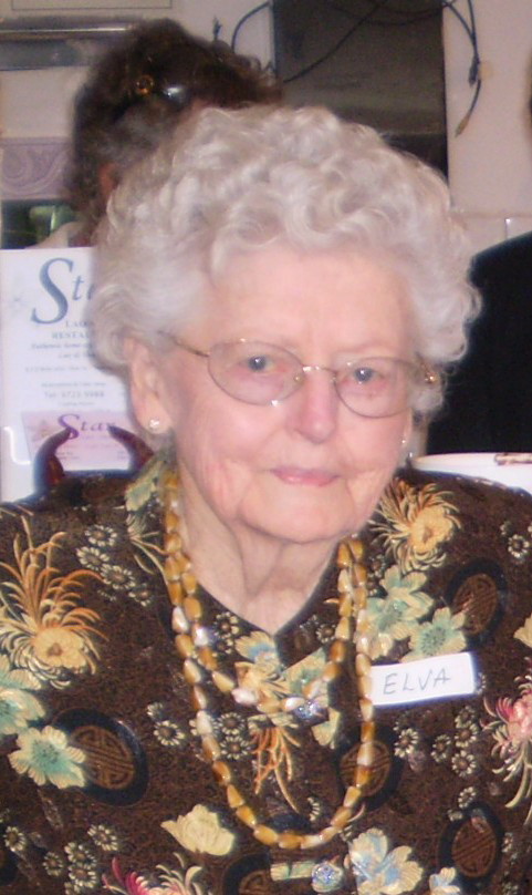 Colour photograph of an elderly woman with white curly hair and rimless spectacles, in a patterned top with beads, wearing a name badge reading ELVA.