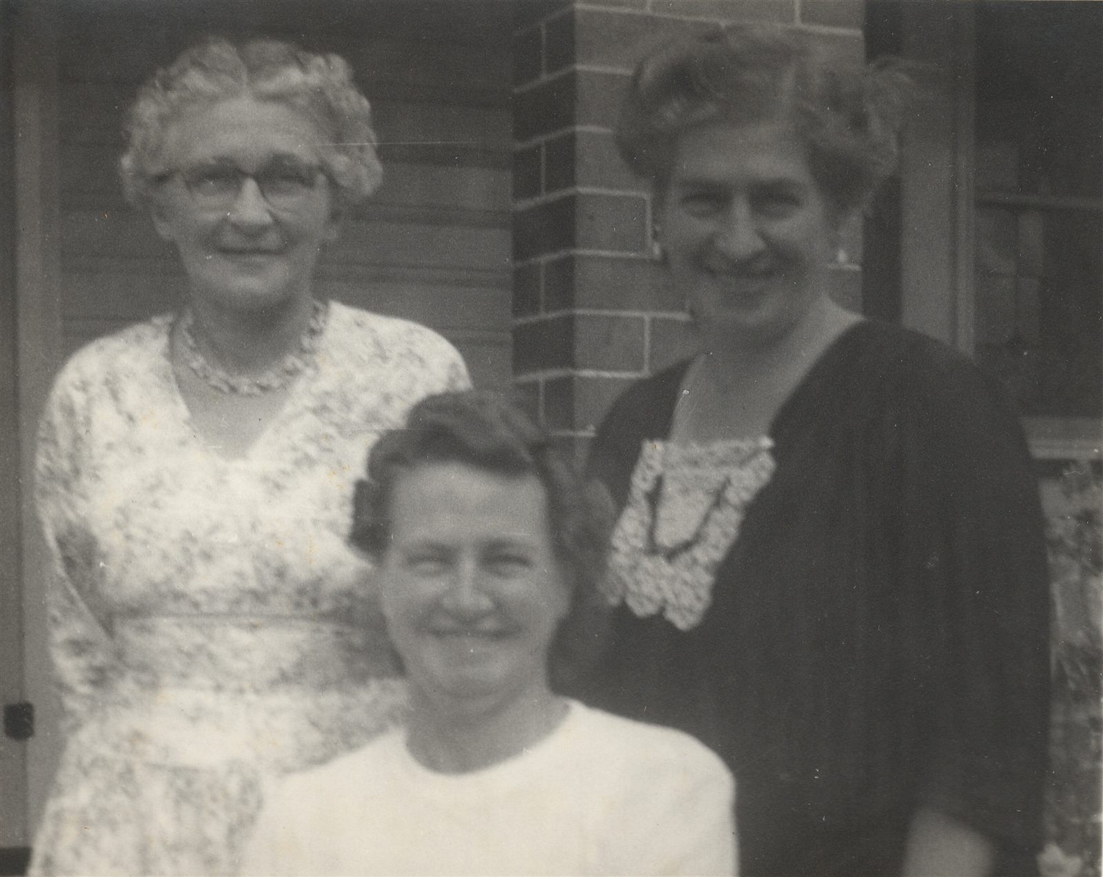 Three older women outside a brick house — one in glasses and a light floral dress, one in front in white, one at right in a dark dress with a lace collar.