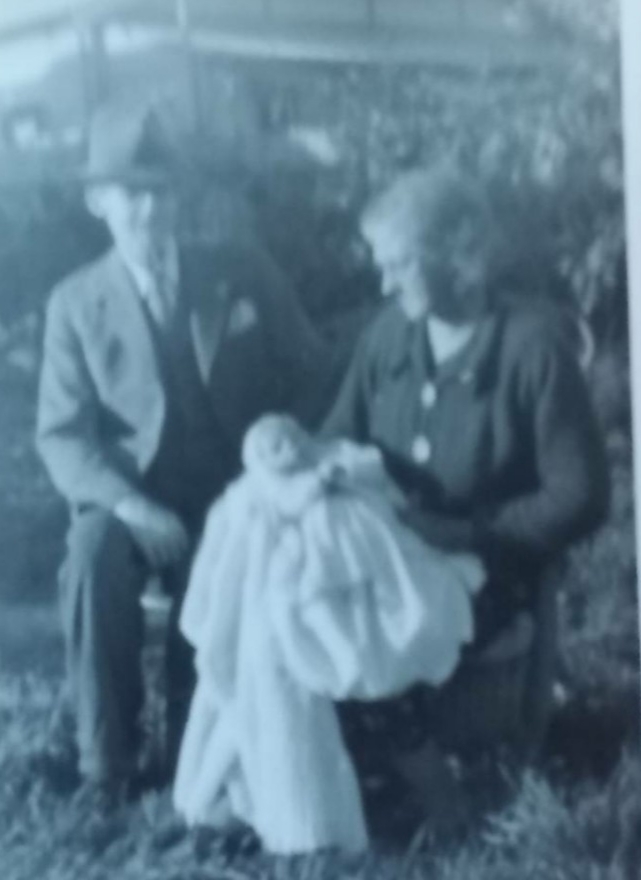 Garden snapshot: a man in a suit and soft felt hat seated on a wooden chair, and a woman in a dark dress standing beside him holding a baby in a long white christening gown.