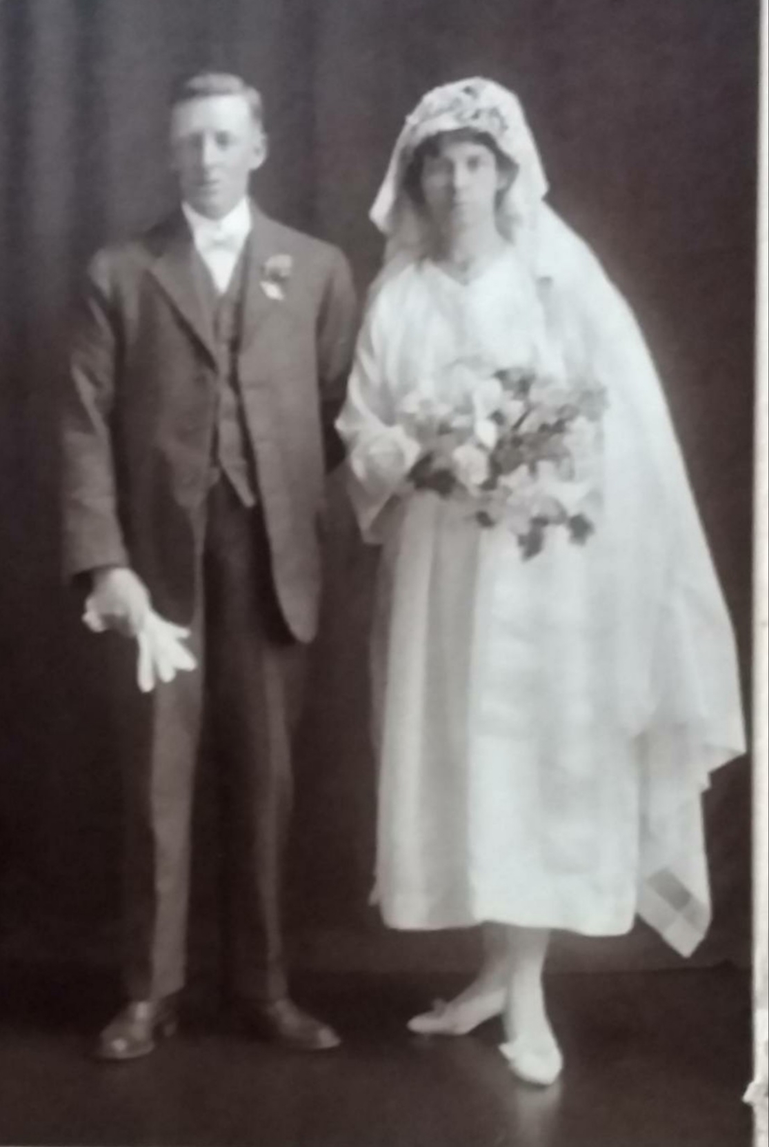 Full-length studio wedding photograph: a man in a light suit holding gloves, and a bride in a lace cap and long veil holding a large trailing bouquet.