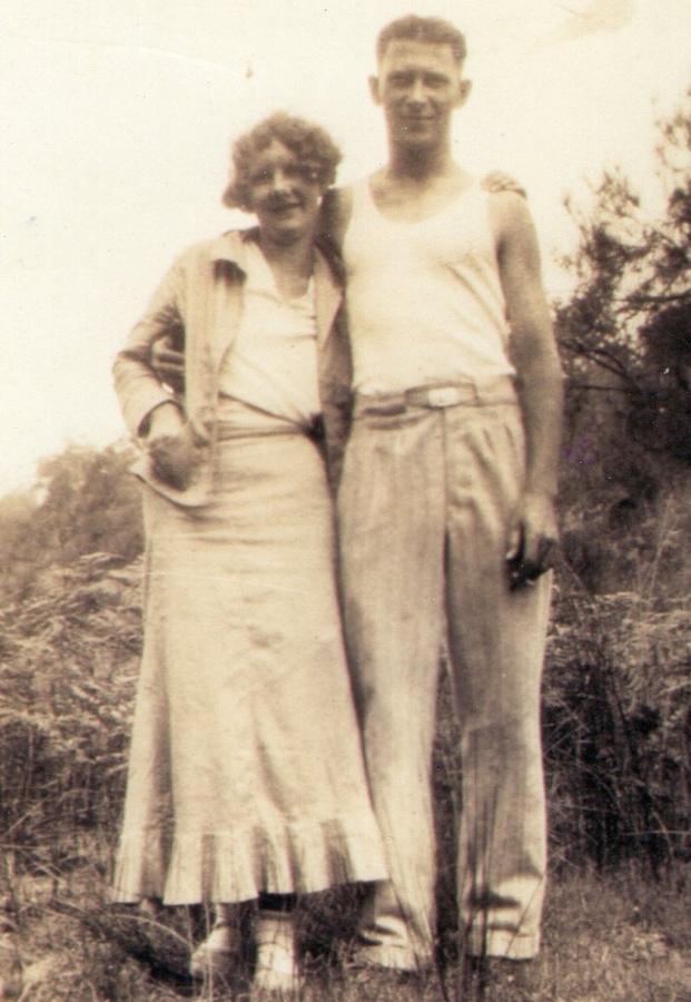 Outdoor snapshot of a young couple standing together, the man in a suit and the woman in a light patterned dress.