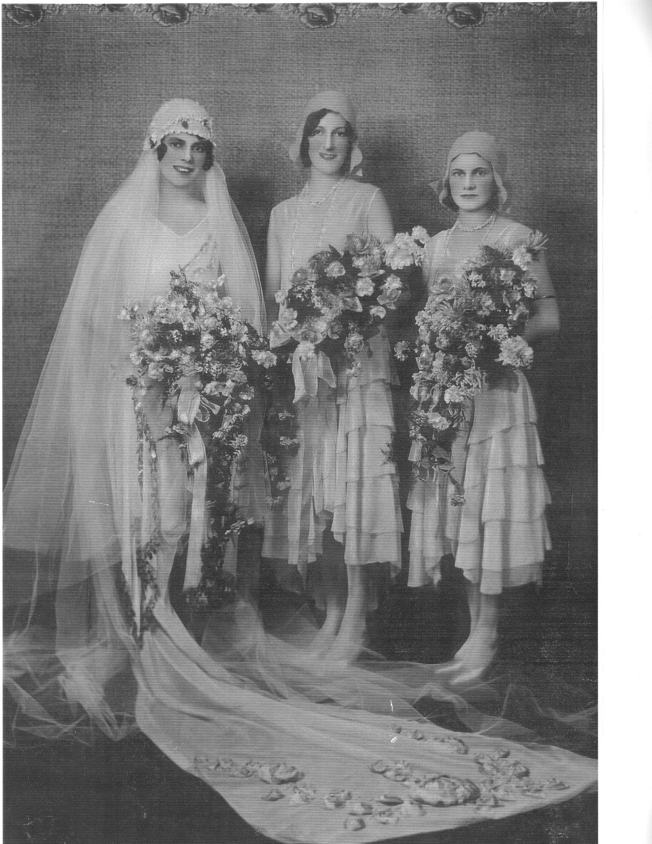 Full-length studio photograph of a bride in a long veil and embroidered train holding a large cascade bouquet, flanked by two attendants in calf-length frocks and close-fitting headdresses, each also holding a bouquet.