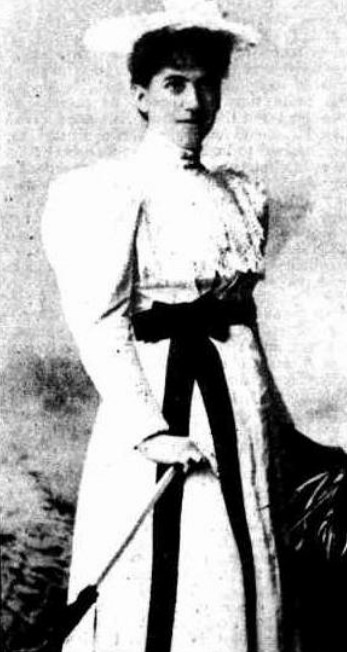 Half-tone newspaper portrait of a young woman standing full-length in a pale high-necked dress with a dark sash and a wide-brimmed hat, holding a long furled parasol angled to the ground.