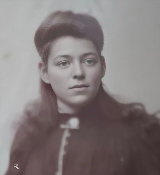 A hand-tinted studio portrait of a young woman with dark hair swept up and back, in a dark fur-trimmed coat, looking a little to one side.