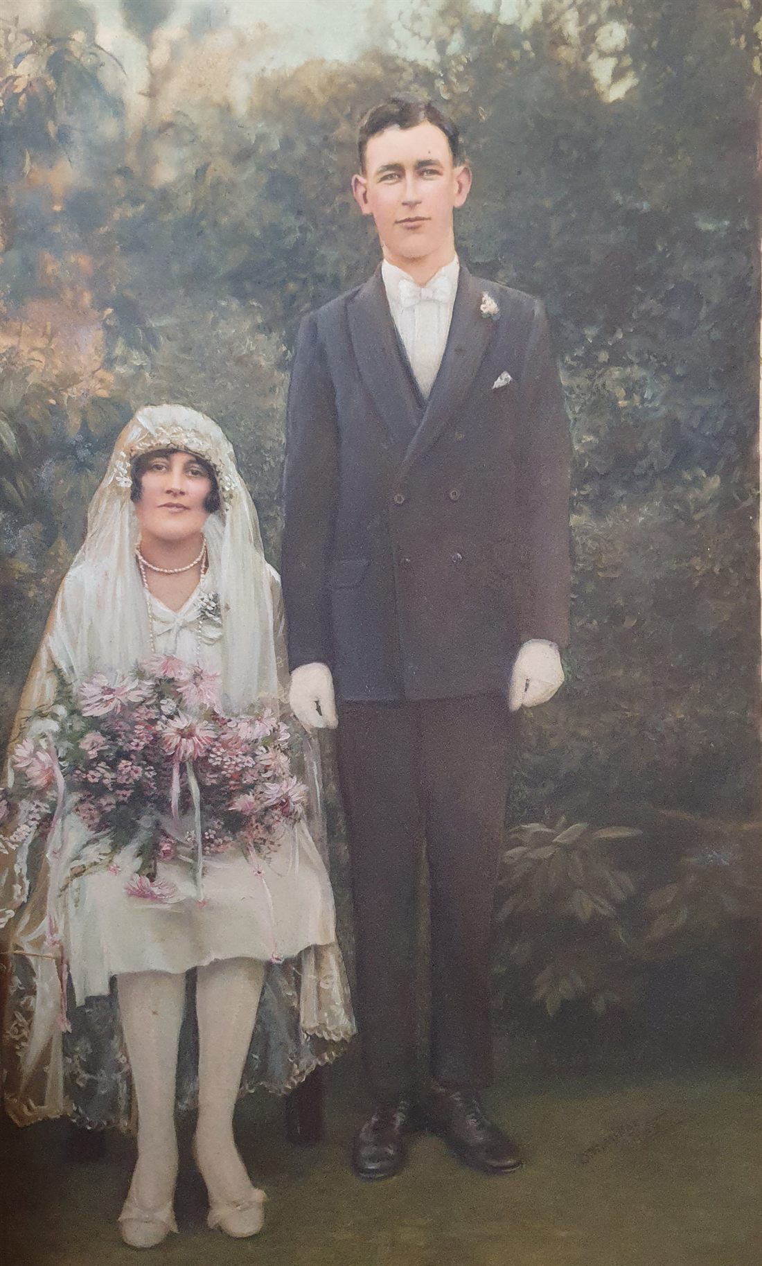 Hand-tinted wedding portrait: a seated bride in a drop-waisted gown and lace veil holding a large trailing bouquet, and a tall standing groom in a double-breasted suit and white bow tie.