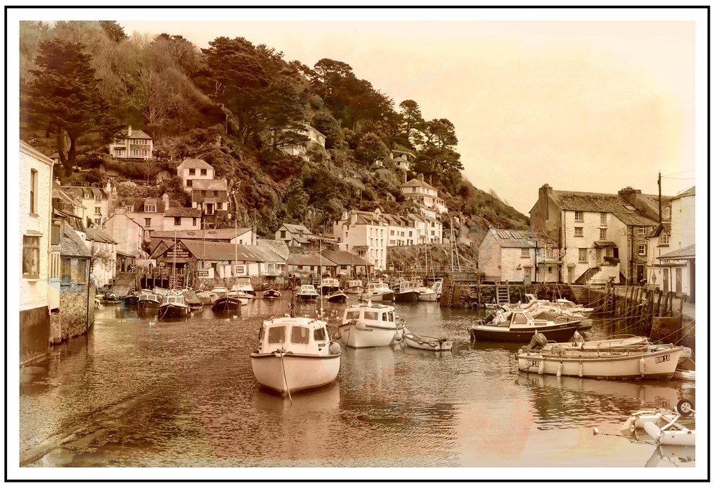 A steep-sided Cornish fishing harbour at low tide — small open boats resting on the mud and moored along the channel, whitewashed cottages stacked up a wooded hillside behind, the whole scene washed in soft sepia and gold like a watercolour.