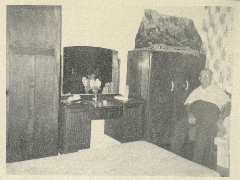 Interior snapshot of a bedroom with a waterfall-front Art Deco dressing table and tallboy; a man in a white short-sleeved shirt and glasses sits at the right.