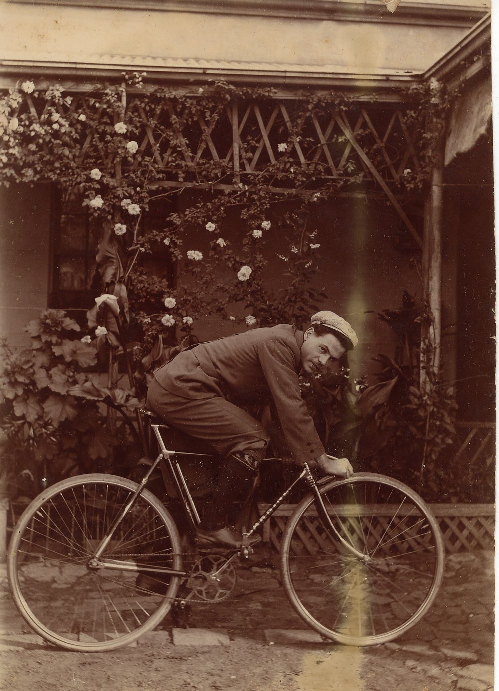 Sepia photograph of a young man with a moustache, in a tweed suit and flat cap, seated on a safety bicycle in front of a verandah covered with climbing roses.