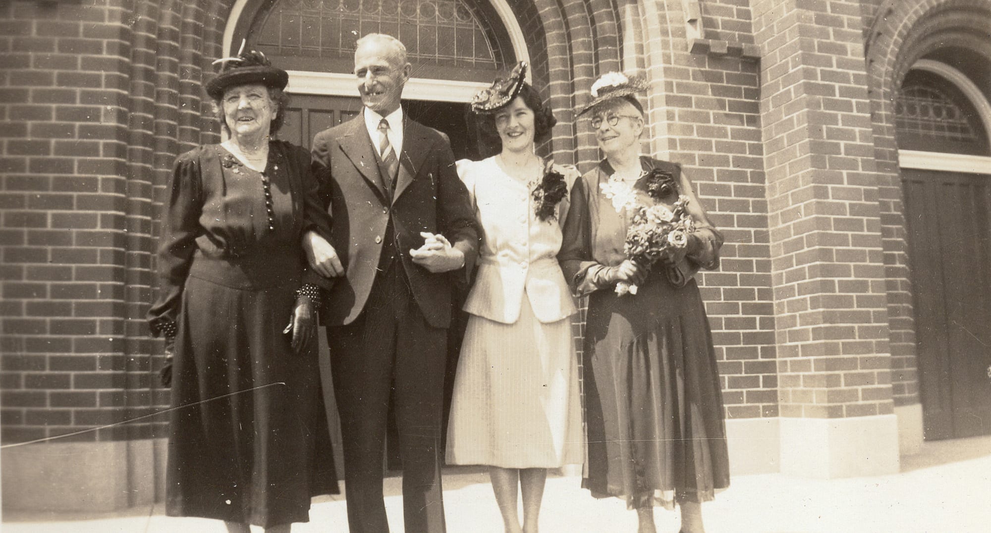 Four people in the sun before a large round-arched brick church doorway: an elderly woman in a dark dress and feathered hat, a balding man in a light suit with a buttonhole, his arm linked with a dark-haired younger woman in a pale peplum suit and flowered hat with a dark corsage, and an elderly bespectacled woman in a dark dress holding a large bouquet.