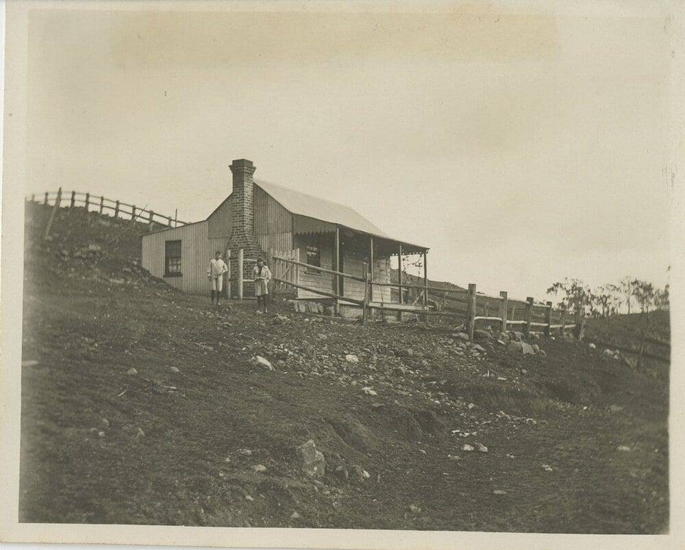 A small slab-and-corrugated-iron cottage with a brick chimney and a skillion verandah, on a bare, eroded hillside with post-and-rail fencing; two children stand by the chimney.