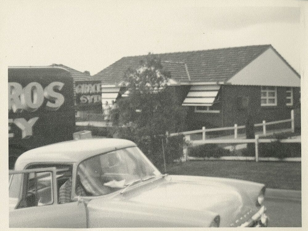 Street snapshot: a large furniture-removal pantechnicon with part-legible lettering parked in front of a brick, tile-roofed house with canvas awnings and a low front fence, a 1950s car in the foreground.
