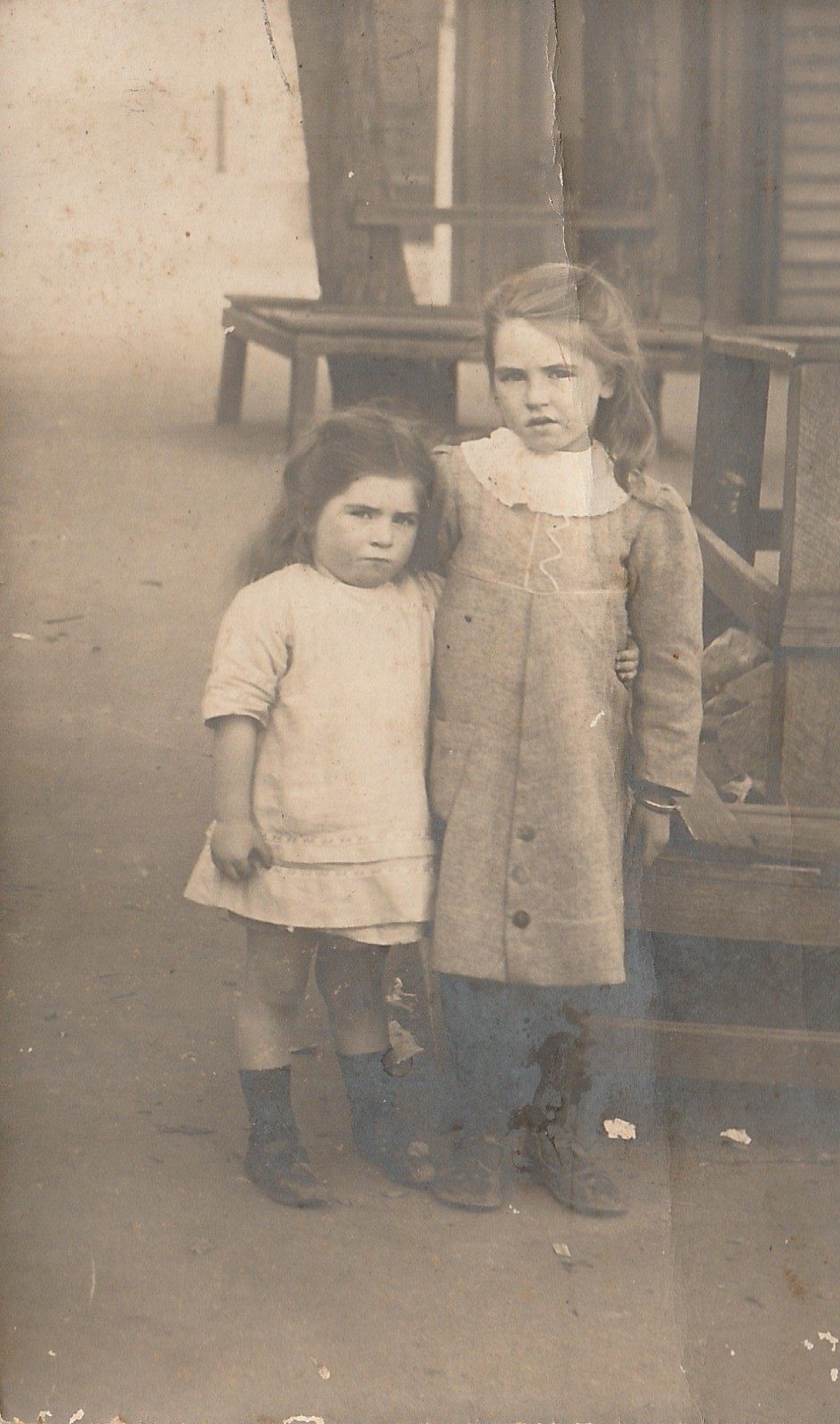 Two young girls stand close together on a verandah — the elder in a buttoned coat with a wide lace collar, the younger in a plain pinafore and boots.