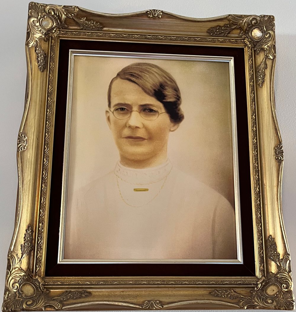 A hand-tinted studio portrait, in an ornate gilt frame, of a woman with dark hair swept to one side, wearing round wire-framed spectacles, a high lace collar and a small gold brooch.