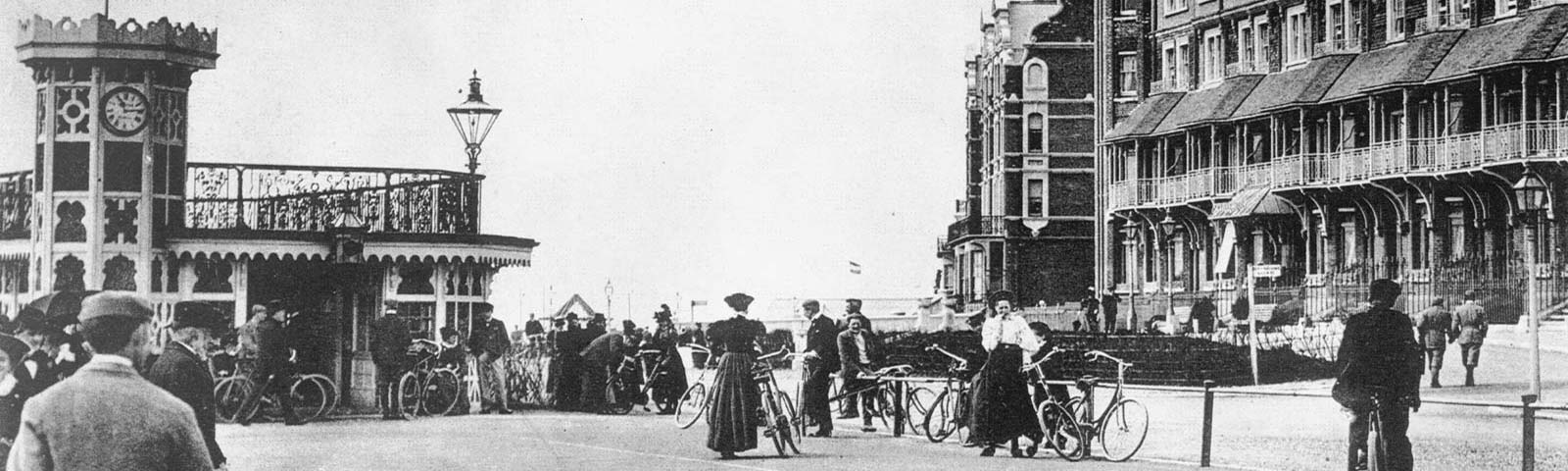 A wide view of an Edwardian seaside promenade — a cast-iron shelter and clock tower at the left, a long balconied hotel terrace at the right, men and women with bicycles walking the front.