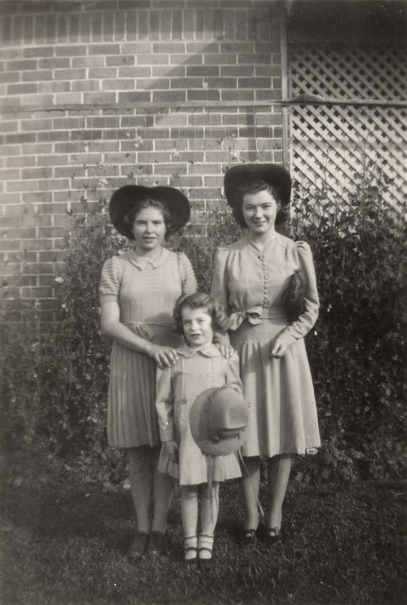 Three girls in front of a brick house wall and a hedge — two older girls in wide-brimmed hats and belted dresses, and a small girl in a light coat holding a round hat.