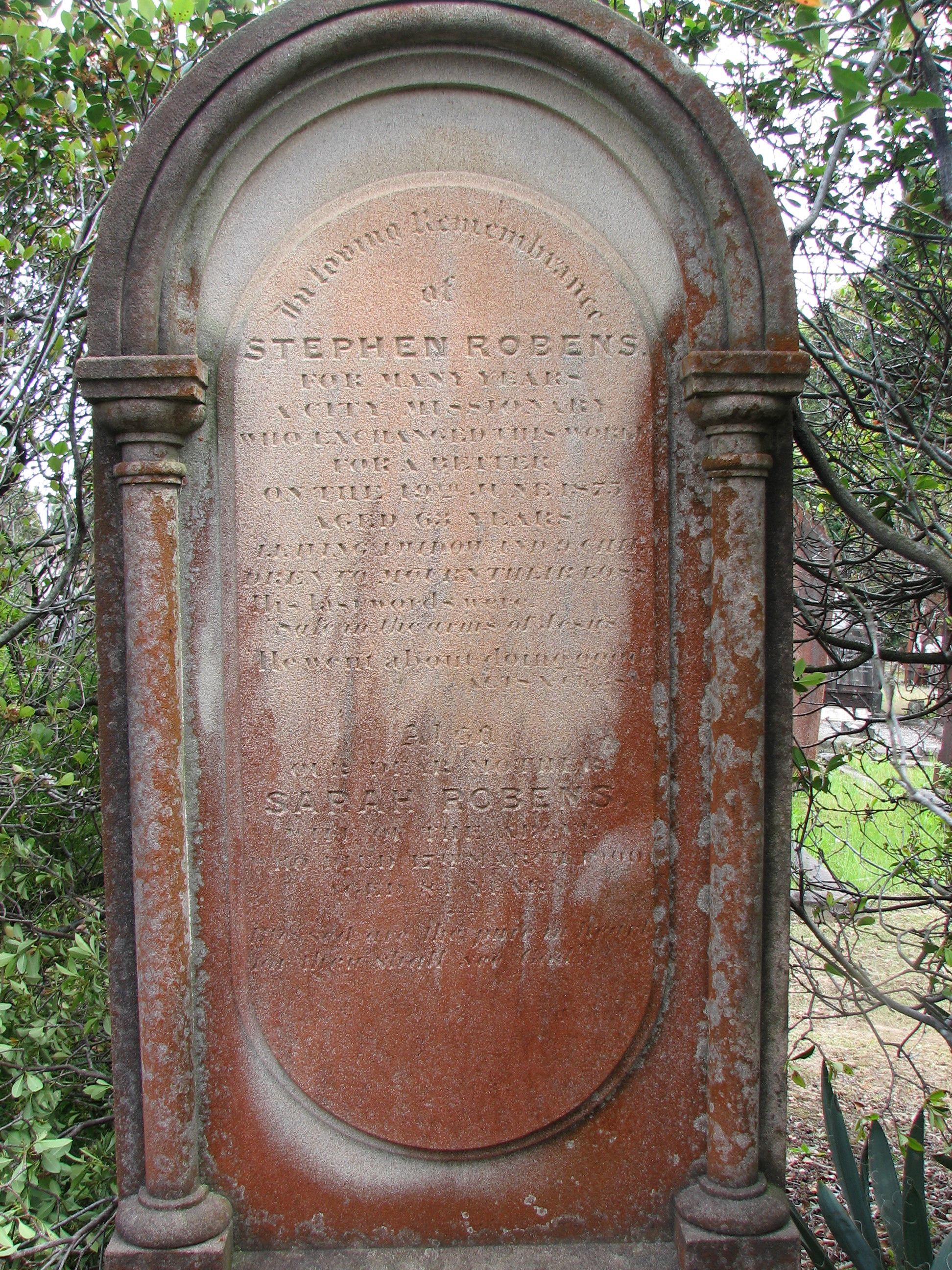A weathered sandstone headstone with an arched top and engaged columns, standing among dark shrubbery, its inscription worn faint.