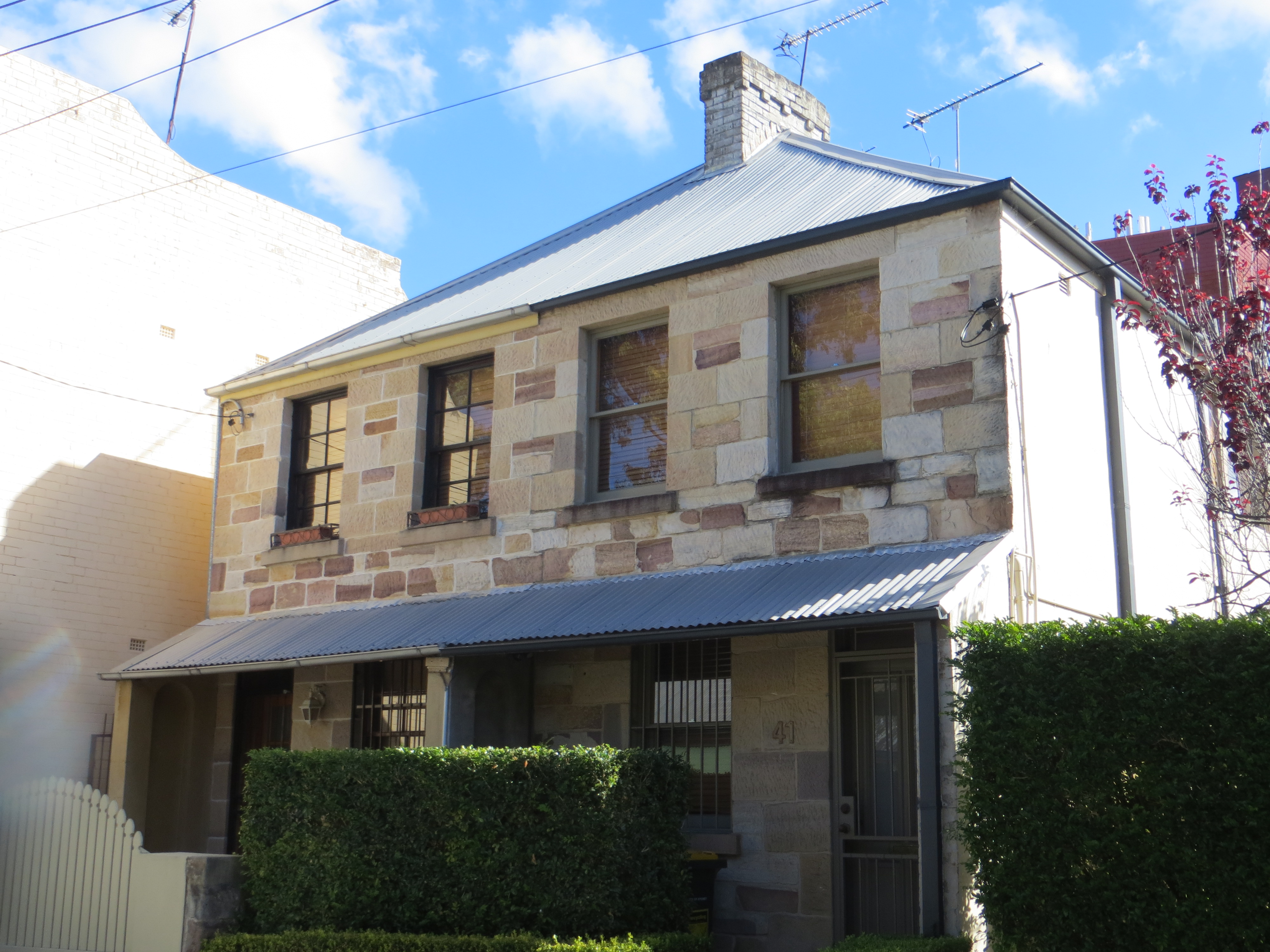 Two joined two-storey sandstone terrace houses with corrugated-iron awnings and hipped roofs, behind low front fences and a clipped hedge, under a blue sky.