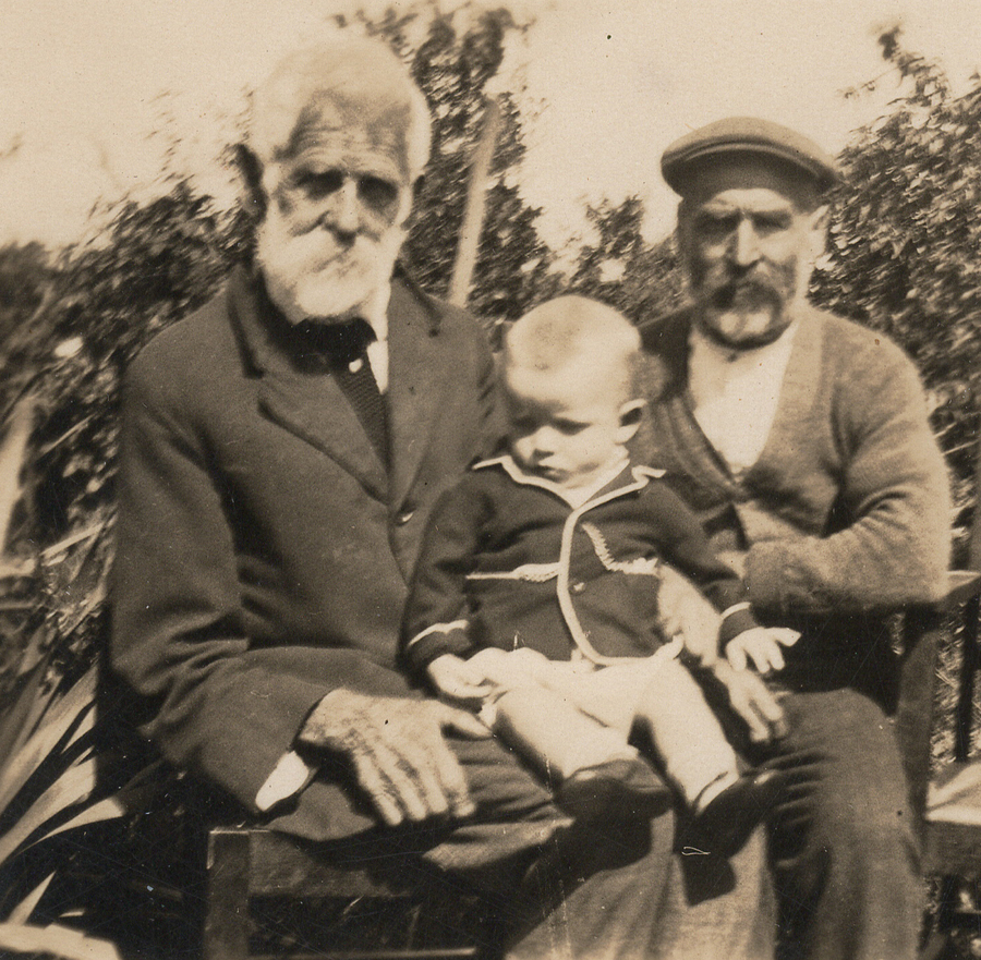 Two elderly men seated outdoors on a garden bench — the white-bearded Thomas Robens on the left, his son Archibald in a flat cap on the right — with an infant in a sailor jacket held between them.